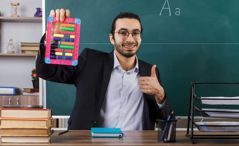 smiling showing thumb up male teacher wearing glasses holding abacus sitting at table with school tools in classroom SSUCv3H4sIAAAAAAAACpyRz27DIAzG75P2DhHnRqJtWpq9SrWD+dMElUIFpNNU9d1nIFScd8M/25/9mefnR9cRDkEL8tU9U4SxNmYJ0UPUziKmm5V7ZaXyLXn4/ZG2QEkdnddgWsghitnCTSG0izEJv3KShAhxCSqk6SsSENWEGgW+JcqK5xJ3NZGT2IEpQjYNCwvPrKIi/q/O8viubmBSVvzmhRsjXhkFxci5lJLrT1T+1lqDRWrXuHo4ASYV7Bulu9dC26lpc3HOZ69twi02+rTCe3NinLsDN+nEF9RUlc8QApbLyptBAj/Z3Zo51sVsYFUlEn8ihdv97rhjbGBbOo4jY6fdWlB+b9aok9epQhiLq5atdZ02IPREGaP80ks+0H6QMPb8AIdebAW/CAbA4YSHf/0BAAD//wMAuaMqXJgCAAA=